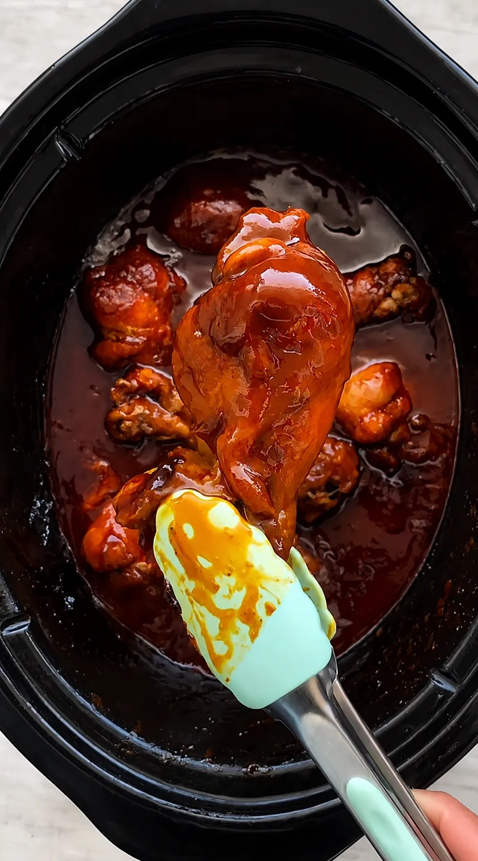 BBQ chicken drumsticks in a black crockpot being basted with sauce